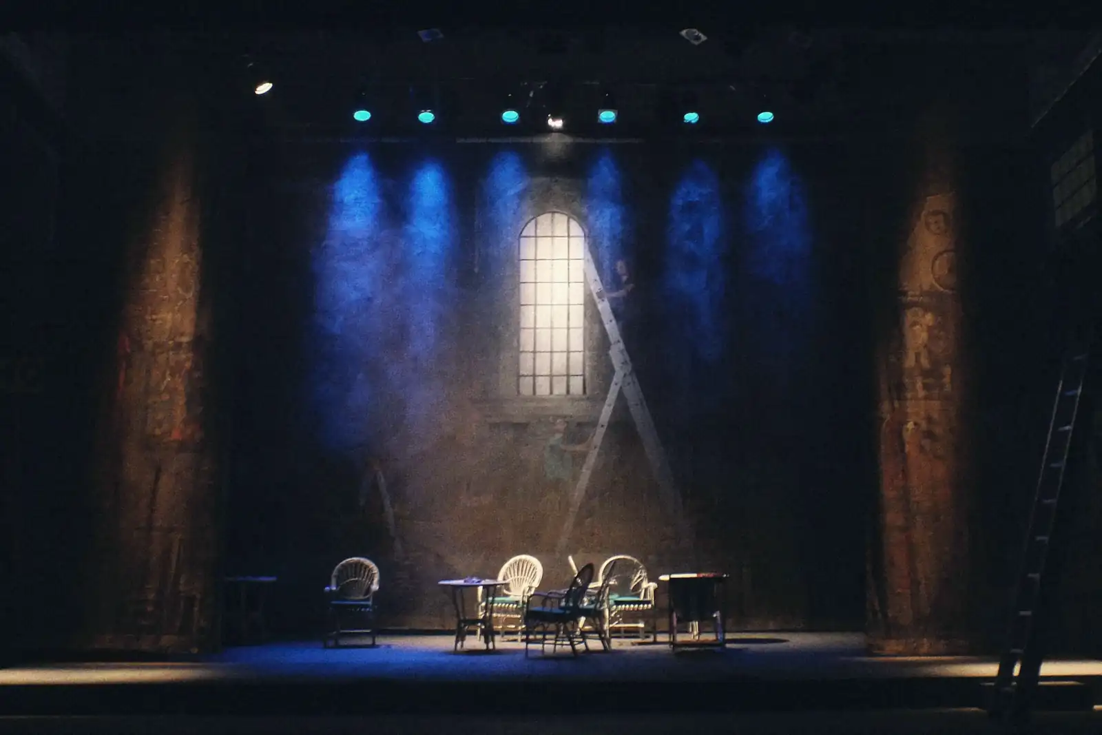 An empty theatre stage in half light: café chairs and small tables under blue spotlights, an arched window painted on the backdrop